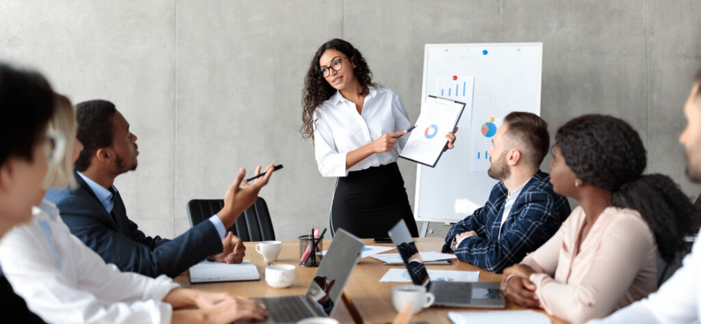 Latin Businesswoman Making Business Presentation For Colleagues Giving Speech On Corporate Meeting Standing Near Blackboard In Modern Office. Female Worker Presenting Business Project